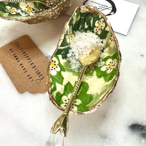May include: Two green and gold oyster shell salt and pepper dishes with white floral designs. Each dish has a gold spoon with a crystal tip. The text "SALT + PEPPER DISHES BY EMMA WADE ART" is on a brown tag.