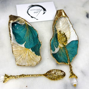 May include: Two decorative oyster shell dishes with gold edging, teal and gold leaf design, and a gold spoon with a crystal handle. The dishes and spoon are on a marble surface. A business card with the text "emma wade art" is in the background.