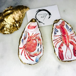 Puede incluir: Conchas de ostra decorativas con bordes dorados y diseños de mariscos. Una concha es dorada. Dos conchas presentan una ilustración de cangrejo y langosta. El nombre del artista, "emma wade art", está en una tarjeta.