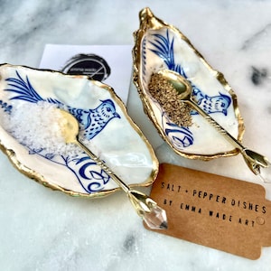 Pode incluir: Dois saleiros e pimenteiros de cerâmica branca em forma de concha de ostra com detalhes dourados e desenhos de pássaros azuis. Cada prato vem com uma colher dourada com um cabo de cristal transparente em forma de ponta de flecha. O texto "SALT + PEPPER DISHES BY EMMA WADE ART" está em uma etiqueta de papel marrom.