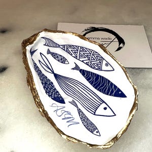 Luxury hand gilded Dorset oyster shell trinket dish featuring a refined blue fish motif, edged in liquid gold leaf and sealed in crystal clear resin. Sustainable coastal ring holder handmade by the artist in Surrey and presented in eco friendly branded gift packaging. Elegant beach house decor piece ideal for jewellery storage, wedding styling or sophisticated coastal interiors.
