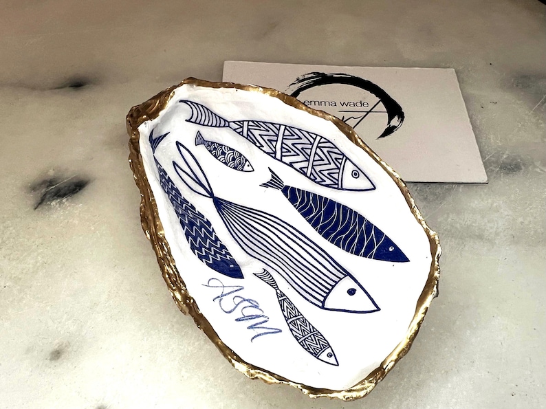 Luxury hand gilded Dorset oyster shell trinket dish featuring a refined blue fish motif, edged in liquid gold leaf and sealed in crystal clear resin. Sustainable coastal ring holder handmade by the artist in Surrey and presented in eco friendly branded gift packaging. Elegant beach house decor piece ideal for jewellery storage, wedding styling or sophisticated coastal interiors.