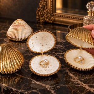 May include: Three decorative seashell-shaped jewelry boxes with metallic gold exteriors and cream-colored interiors. Two boxes are open, each containing a gold ring with a single white pearl and a clear stone. One box is closed, showing a ribbed gold shell.