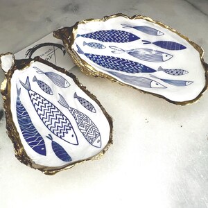 Luxury hand gilded Dorset oyster shell trinket dish featuring a refined blue fish motif, edged in liquid gold leaf and sealed in crystal clear resin. Sustainable coastal ring holder handmade by the artist in Surrey and presented in eco friendly branded gift packaging. Elegant beach house decor piece ideal for jewellery storage, wedding styling or sophisticated coastal interiors.