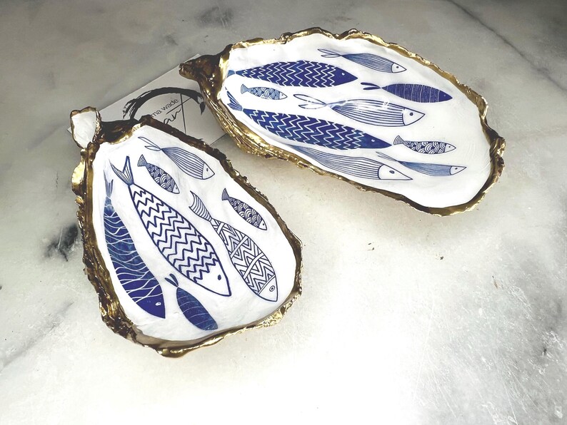 Luxury hand gilded Dorset oyster shell trinket dish featuring a refined blue fish motif, edged in liquid gold leaf and sealed in crystal clear resin. Sustainable coastal ring holder handmade by the artist in Surrey and presented in eco friendly branded gift packaging. Elegant beach house decor piece ideal for jewellery storage, wedding styling or sophisticated coastal interiors.
