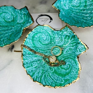 May include: Three decorative teal blue and gold shell-shaped trinket dishes. The centre dish has a gold watch and bracelet inside.
