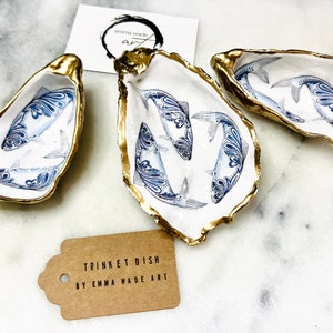 Peut inclure: Trois coupelles à bijoux en forme de coquille d'huître blanche avec des bords dorés. Chaque coupelle présente une illustration bleue et blanche d'un poisson. Les coupelles sont posées sur une surface en marbre blanc. Le texte "TRINKET DISH BY EMMA WADE ART" est sur une étiquette brune.