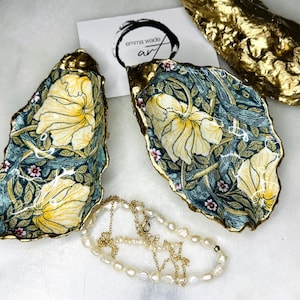 May include: Two gold oyster shell dishes with a floral pattern in shades of green, yellow, and pink. The shells are decorated with a gold trim. A pearl necklace is lying on a white surface.