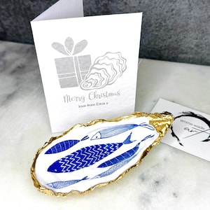 Luxury hand gilded Dorset oyster shell trinket dish featuring a refined blue fish motif, edged in liquid gold leaf and sealed in crystal clear resin. Sustainable coastal ring holder handmade by the artist in Surrey and presented in eco friendly branded gift packaging. Elegant beach house decor piece ideal for jewellery storage, wedding styling or sophisticated coastal interiors.