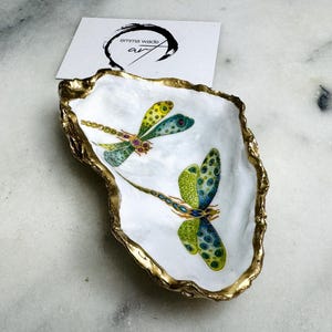 Handmade dragonfly oyster shell trinket dish with hand-gilded gold leaf edges and glossy resin finish, created from sustainably sourced Dorset shells. Unique coastal jewellery holder and ring dish, ideal as a vanity tray, trinket tray or decorative keepsake. Nature inspired home decor gift for birthdays, weddings, anniversaries, housewarmings and Mother’s Day. One of a kind artisan shell dish, beautifully presented in a gift box.