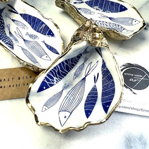 Hand-gilded Dorset oyster shell trinket dish featuring a blue fish motif, edged with liquid gold leaf and sealed in high gloss resin. Coastal ring holder and nautical home decor piece displayed with multiple shells. Sustainable oyster shell art handmade in Surrey, ideal as a jewellery dish, wedding favour, beach house accessory or ocean lover gift.