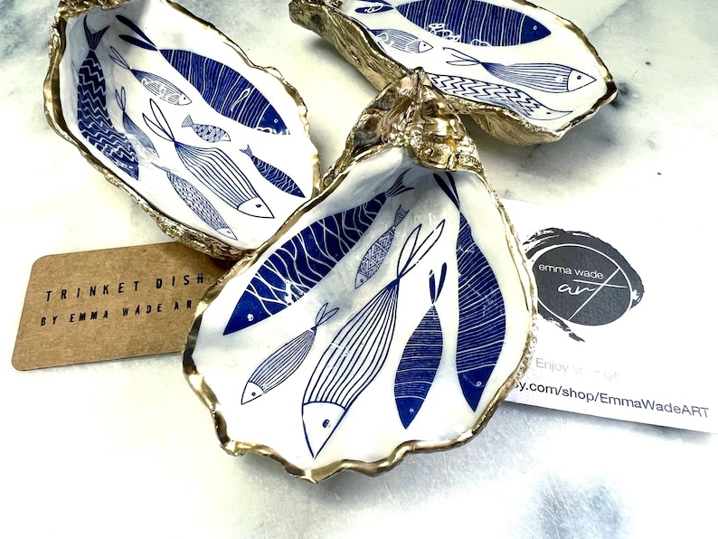 Hand-gilded Dorset oyster shell trinket dish featuring a blue fish motif, edged with liquid gold leaf and sealed in high gloss resin. Coastal ring holder and nautical home decor piece displayed with multiple shells. Sustainable oyster shell art handmade in Surrey, ideal as a jewellery dish, wedding favour, beach house accessory or ocean lover gift.