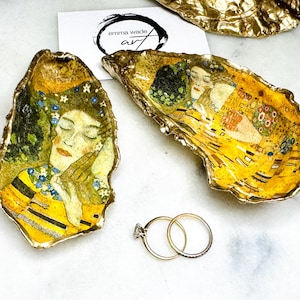Op de afbeelding: Twee decoratieve oesterschelpen met gouden randen, met kunstwerken geïnspireerd op Gustav Klimt. Eén schelp toont een vrouwenportret, de andere een paar. Er zijn ook twee gouden ringen aanwezig. Het logo van de kunstenaar, "emma wade art", is zichtbaar.