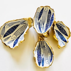 Luxury hand gilded Dorset oyster shell trinket dish featuring a refined blue fish motif, edged in liquid gold leaf and sealed in crystal clear resin. Sustainable coastal ring holder handmade by the artist in Surrey and presented in eco friendly branded gift packaging. Elegant beach house decor piece ideal for jewellery storage, wedding styling or sophisticated coastal interiors.