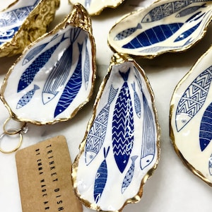 Hand-gilded Dorset oyster shell trinket dish featuring a blue fish motif, edged with liquid gold leaf and sealed in high gloss resin. Coastal ring holder and nautical home decor piece displayed with multiple shells. Sustainable oyster shell art handmade in Surrey, ideal as a jewellery dish, wedding favour, beach house accessory or ocean lover gift.