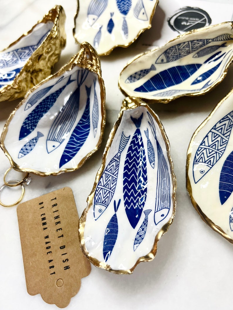 Hand-gilded Dorset oyster shell trinket dish featuring a blue fish motif, edged with liquid gold leaf and sealed in high gloss resin. Coastal ring holder and nautical home decor piece displayed with multiple shells. Sustainable oyster shell art handmade in Surrey, ideal as a jewellery dish, wedding favour, beach house accessory or ocean lover gift.
