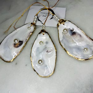 May include: Three gold-rimmed oyster shell ornaments with white pearls inside. Each ornament has a gold string attached.