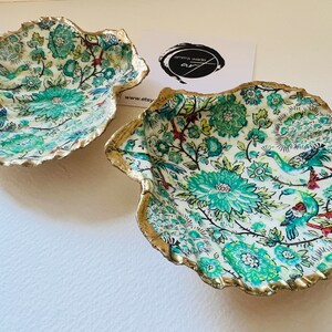 May include: Two decorative shell-shaped bowls with a white background and a floral pattern in shades of green, blue, and red. The bowls have a gold-painted rim.
