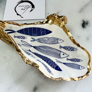 Hand-gilded Dorset oyster shell trinket dish featuring a blue fish motif, edged with liquid gold leaf and sealed in high gloss resin. Coastal ring holder and nautical home decor piece displayed with multiple shells. Sustainable oyster shell art handmade in Surrey, ideal as a jewellery dish, wedding favour, beach house accessory or ocean lover gift.