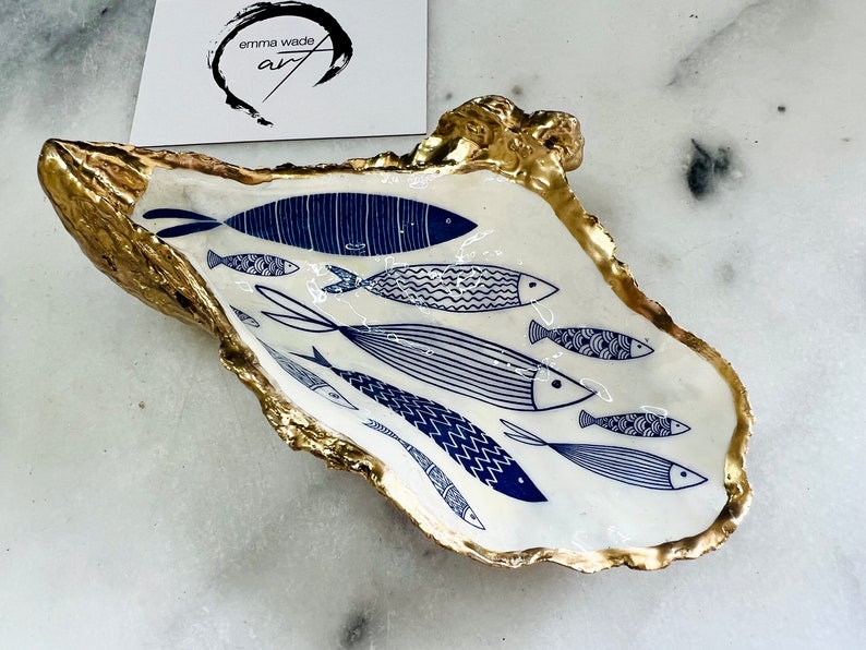 Hand-gilded Dorset oyster shell trinket dish featuring a blue fish motif, edged with liquid gold leaf and sealed in high gloss resin. Coastal ring holder and nautical home decor piece displayed with multiple shells. Sustainable oyster shell art handmade in Surrey, ideal as a jewellery dish, wedding favour, beach house accessory or ocean lover gift.