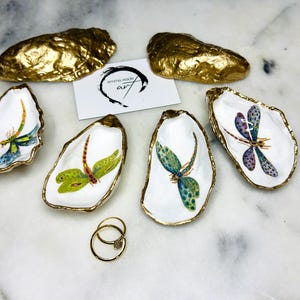 Handmade dragonfly oyster shell trinket dish with hand-gilded gold leaf edges and glossy resin finish, created from sustainably sourced Dorset shells. Unique coastal jewellery holder and ring dish, ideal as a vanity tray, trinket tray or decorative keepsake. Nature inspired home decor gift for birthdays, weddings, anniversaries, housewarmings and Mother’s Day. One of a kind artisan shell dish, beautifully presented in a gift box.