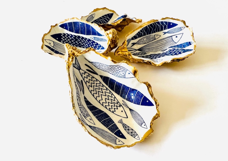 Luxury hand gilded Dorset oyster shell trinket dish featuring a refined blue fish motif, edged in liquid gold leaf and sealed in crystal clear resin. Sustainable coastal ring holder handmade by the artist in Surrey and presented in eco friendly branded gift packaging. Elegant beach house decor piece ideal for jewellery storage, wedding styling or sophisticated coastal interiors.