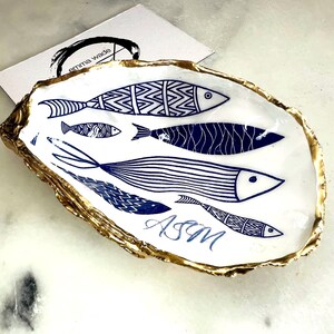 Luxury hand gilded Dorset oyster shell trinket dish featuring a refined blue fish motif, edged in liquid gold leaf and sealed in crystal clear resin. Sustainable coastal ring holder handmade by the artist in Surrey and presented in eco friendly branded gift packaging. Elegant beach house decor piece ideal for jewellery storage, wedding styling or sophisticated coastal interiors. Personalised.