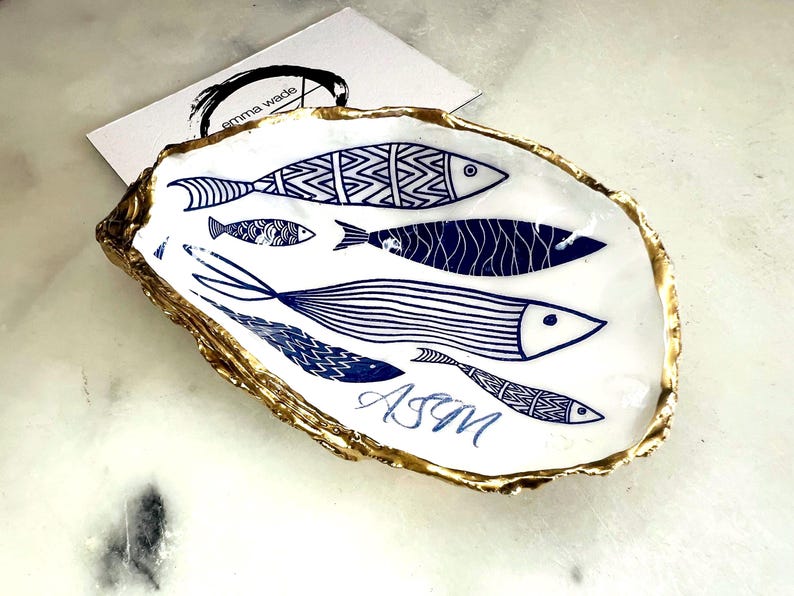 Luxury hand gilded Dorset oyster shell trinket dish featuring a refined blue fish motif, edged in liquid gold leaf and sealed in crystal clear resin. Sustainable coastal ring holder handmade by the artist in Surrey and presented in eco friendly branded gift packaging. Elegant beach house decor piece ideal for jewellery storage, wedding styling or sophisticated coastal interiors. Personalised.
