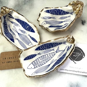 Hand-gilded Dorset oyster shell trinket dish featuring a blue fish motif, edged with liquid gold leaf and sealed in high gloss resin. Coastal ring holder and nautical home decor piece displayed with multiple shells. Sustainable oyster shell art handmade in Surrey, ideal as a jewellery dish, wedding favour, beach house accessory or ocean lover gift.