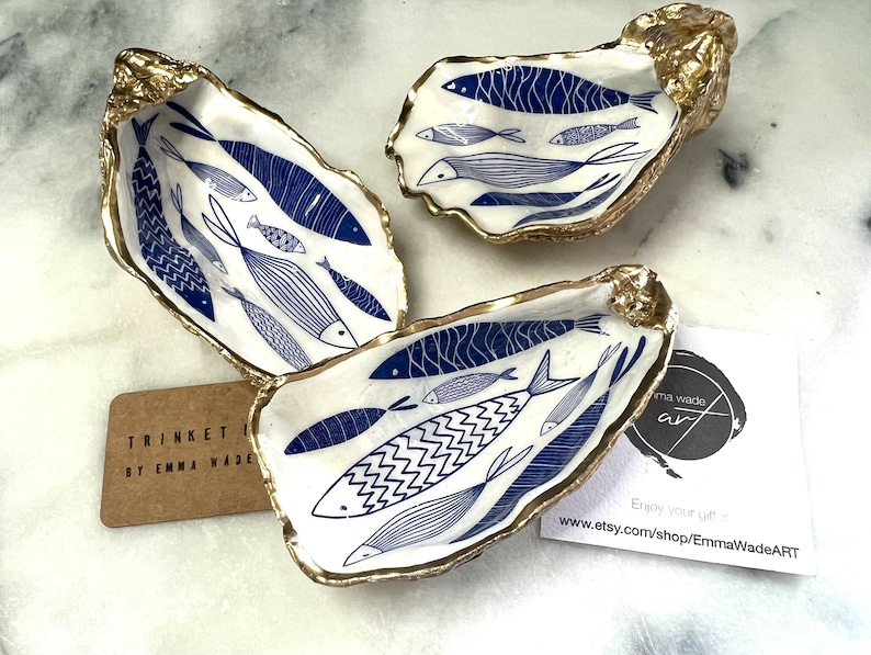 Hand-gilded Dorset oyster shell trinket dish featuring a blue fish motif, edged with liquid gold leaf and sealed in high gloss resin. Coastal ring holder and nautical home decor piece displayed with multiple shells. Sustainable oyster shell art handmade in Surrey, ideal as a jewellery dish, wedding favour, beach house accessory or ocean lover gift.