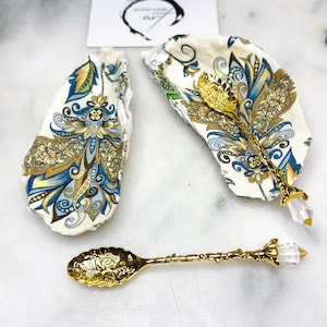 May include: Decorative oyster shell spoon rests with a gold spoon. The shells are painted with a blue, gold, and white floral design. The gold spoon has a crystal accent on the handle. The spoon rests are on a marble surface.