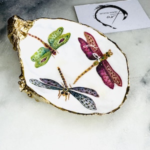 Handmade dragonfly oyster shell trinket dish with hand-gilded gold leaf edges and glossy resin finish, created from sustainably sourced Dorset shells. Unique coastal jewellery holder and ring dish, ideal as a vanity tray, trinket tray or decorative keepsake. Nature inspired home decor gift for birthdays, weddings, anniversaries, housewarmings and Mother’s Day. One of a kind artisan shell dish, beautifully presented in a gift box.