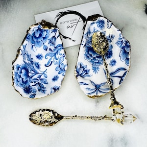 May include: Two blue and white porcelain oyster shell dishes with gold trim. A gold spoon with a clear crystal handle sits in one of the dishes. The spoon is decorated with a floral design.