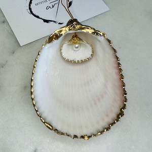 Może przedstawiać: Biała muszla z pozłacanym brzegiem i małą białą perłą w środku. Muszla jest ozdobą.