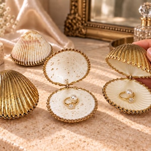 May include: A collection of hinged seashell trinket boxes featuring metallic gold exteriors with ribbed textures. The interiors are finished in a smooth cream colour, with some featuring gold speckles. Each open shell contains a gold ring with a pearl.