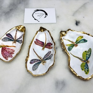 Handmade dragonfly oyster shell trinket dish with hand-gilded gold leaf edges and glossy resin finish, created from sustainably sourced Dorset shells. Unique coastal jewellery holder and ring dish, ideal as a vanity tray, trinket tray or decorative keepsake. Nature inspired home decor gift for birthdays, weddings, anniversaries, housewarmings and Mother’s Day. One of a kind artisan shell dish, beautifully presented in a gift box.