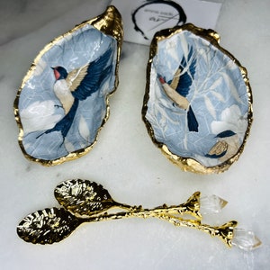 May include: Two gold-rimmed oyster shells with a blue and white floral design and a bird painted on each. The shells are displayed on a white surface with a pair of gold spoons with crystal flower accents.