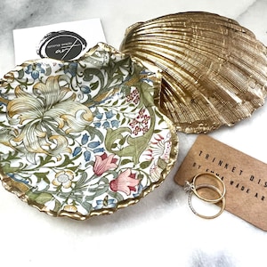 May include: A gold shell trinket dish with a floral pattern. The dish is filled with two gold rings. The dish is labelled "Trinket Dish by Emma Wade Art".