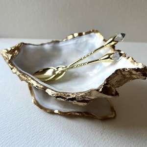 May include: An oyster shell-shaped dish with a gold-painted rim and base. Two gold-coloured spoons with crystal-like accents are inside the white shell. The dish is designed for serving or display, a decorative item.