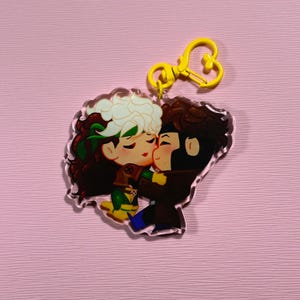 Pode incluir: Um chaveiro de acrílico transparente com um casal de desenhos animados se beijando. A mulher tem cabelo verde e está vestindo uma roupa verde e amarela. O homem tem cabelo castanho e está vestindo uma jaqueta marrom. O chaveiro tem um anel amarelo em forma de coração.