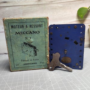 May include: Vintage Meccano toy set with a blue metal plate, a key, and a green box. The box reads "MOTEUR A RESSORT MECCANO N° 1 Fabriqué en France par MECCANO Paris". The set is likely from the mid-20th century.