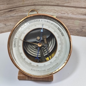 May include: A vintage brass barometer with a white face and black markings. The barometer has a circular dial with a needle that points to the current atmospheric pressure. The dial is labeled with the words "Tempête", "Coé Pluie", "Pluie", "Variable", "Beau Temps", "Fixe", "Très Sec". The barometer is mounted on a wooden base.