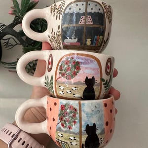 May include: Three stacked ceramic mugs with whimsical, hand-painted designs. Each mug features a different scene: a window with books, a cat overlooking a field, and a cat by a window with birds. The mugs have pink handles and are decorated with various colors.
