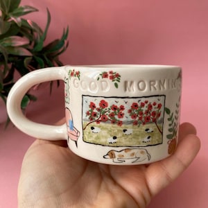Puede incluir: Taza de cerámica blanca con un diseño pintado a mano que presenta un campo de flores rojas, ovejas, un perro y un sofá. La taza tiene las palabras "GOOD MORNING" en relieve en el lateral.