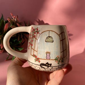 Puede incluir: Una taza de cerámica blanca con un diseño pintado a mano de una ventana con un gato negro sentado en un borde de ladrillo. La ventana tiene un fondo azul claro con una maceta rosa y una pantalla de lámpara verde claro colgando del techo. La taza tiene una asa blanca y está decorada con flores rojas.