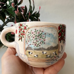 May include: A beige ceramic mug with a hand-painted landscape scene featuring a tree with pink blossoms, sheep, and mountains. The mug has a rounded handle and is decorated with red flowers.