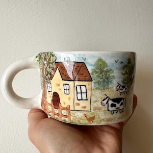 Könnte beinhalten: Ein Keramikbecher mit einer handgemalten Szene, die ein gelbes Haus mit braunem Dach, einen Holzzaun, einen Baum mit rosa Blüten, grüne Bäume, ein kleines braunes Huhn und zwei schwarz-weiße Kühe auf einer Weide unter blauem Himmel zeigt.
