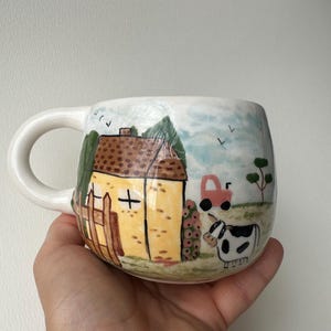 Puede incluir: Una taza de cerámica blanca con una escena rural pintada a mano. El diseño incluye una casa amarilla con techo marrón, una valla de madera, un tractor rosa y una vaca blanca y negra sobre hierba verde bajo un cielo azul claro con aves.