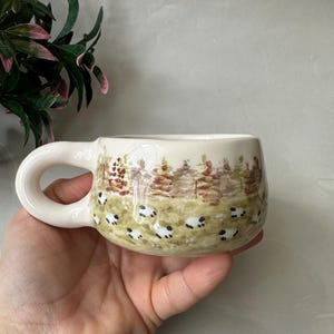 Puede incluir: Una taza de cerámica color crema con asa redondeada, que presenta una escena pintada a mano de un pasto verde lleno de pequeñas ovejas blancas y negras, sobre un fondo de árboles marrones y verdes bajo un cielo de tonos neutros.