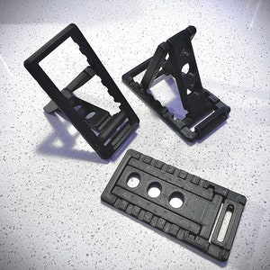 Puede incluir: Tres soportes para teléfono negros, impresos en 3D. Dos están en posición vertical, con un diseño dentado para ajustar el ángulo de visión. El tercero es una base plana con tres agujeros circulares y una ranura rectangular. Todos son de material negro mate.